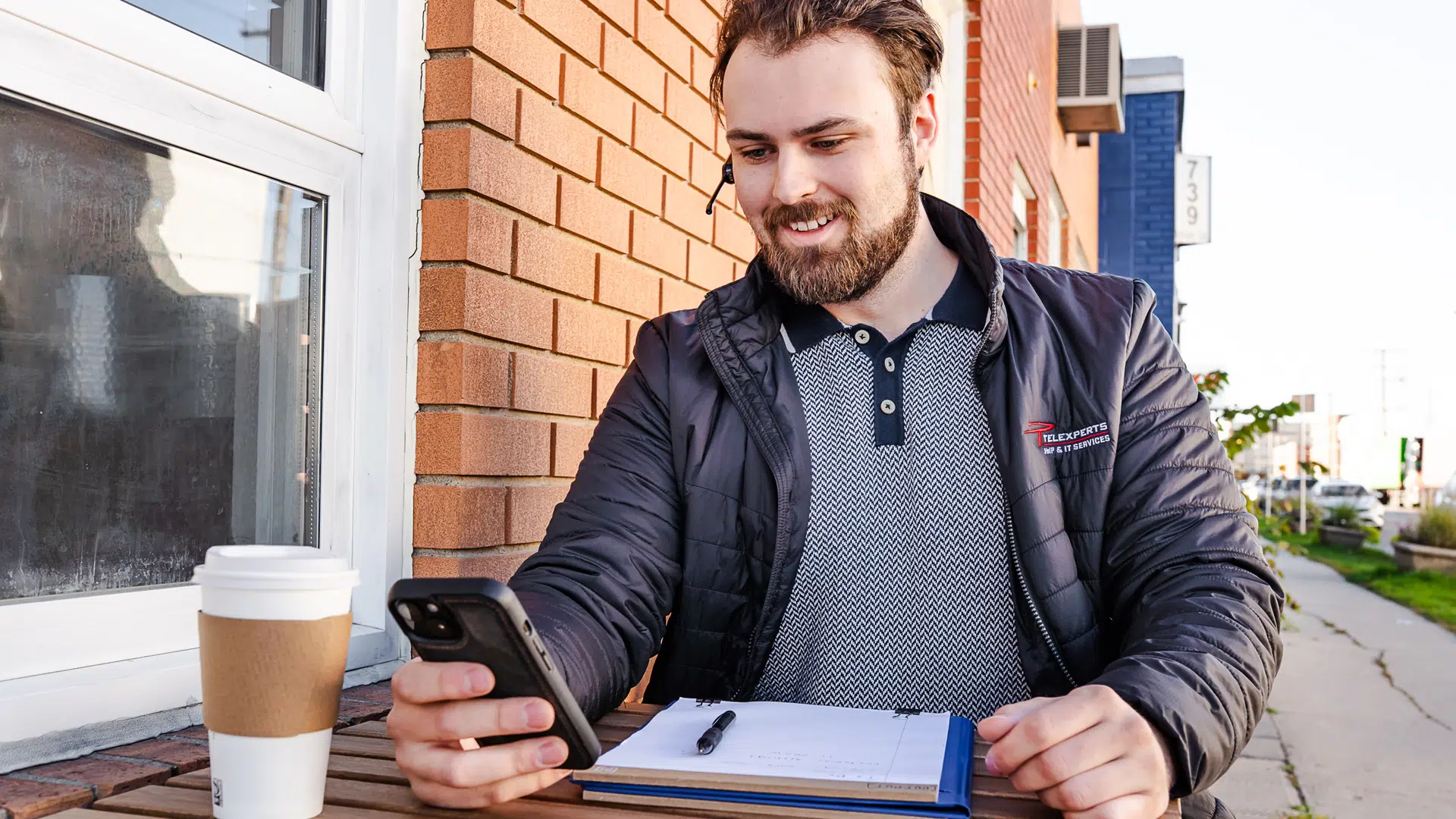 A Telexperts employee using VoIP systems while sitting outside at a table with a notepad in front of him and a cell phone in hand.