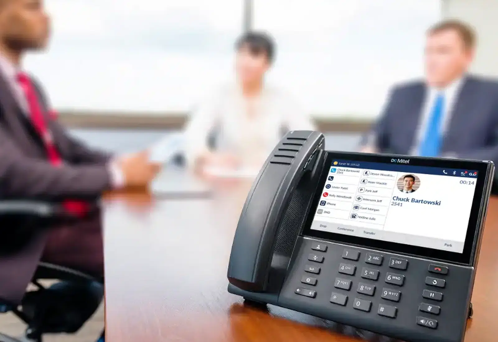 A close up of a Mitel IP phone running software for a conference call.  In the background, three employees are sitting around a table.