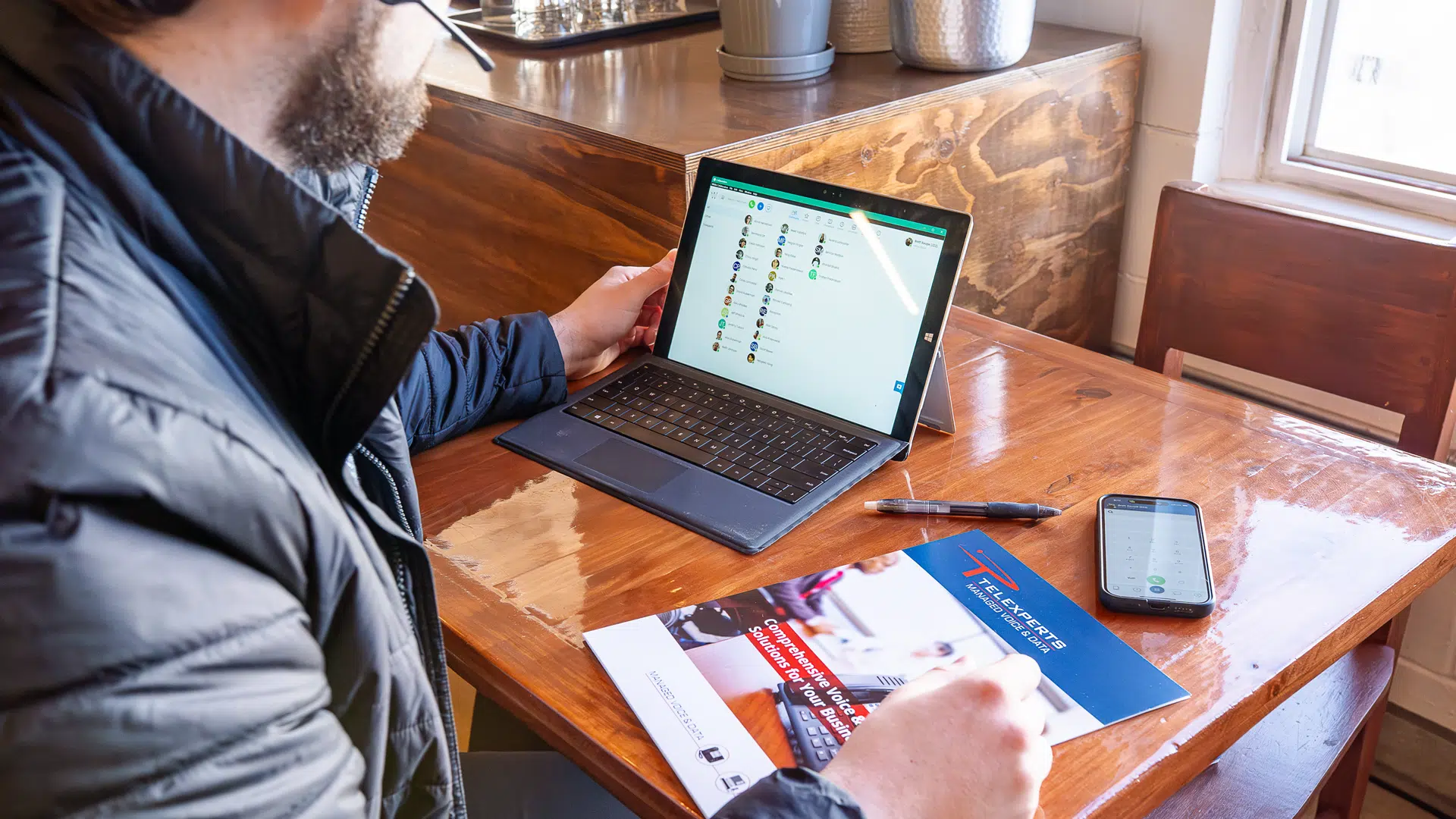 A Telexperts employee sitting at a table with a laptop and smartphone as well as a brochure. The electronics are using VoIP systems to conduct a voice call.