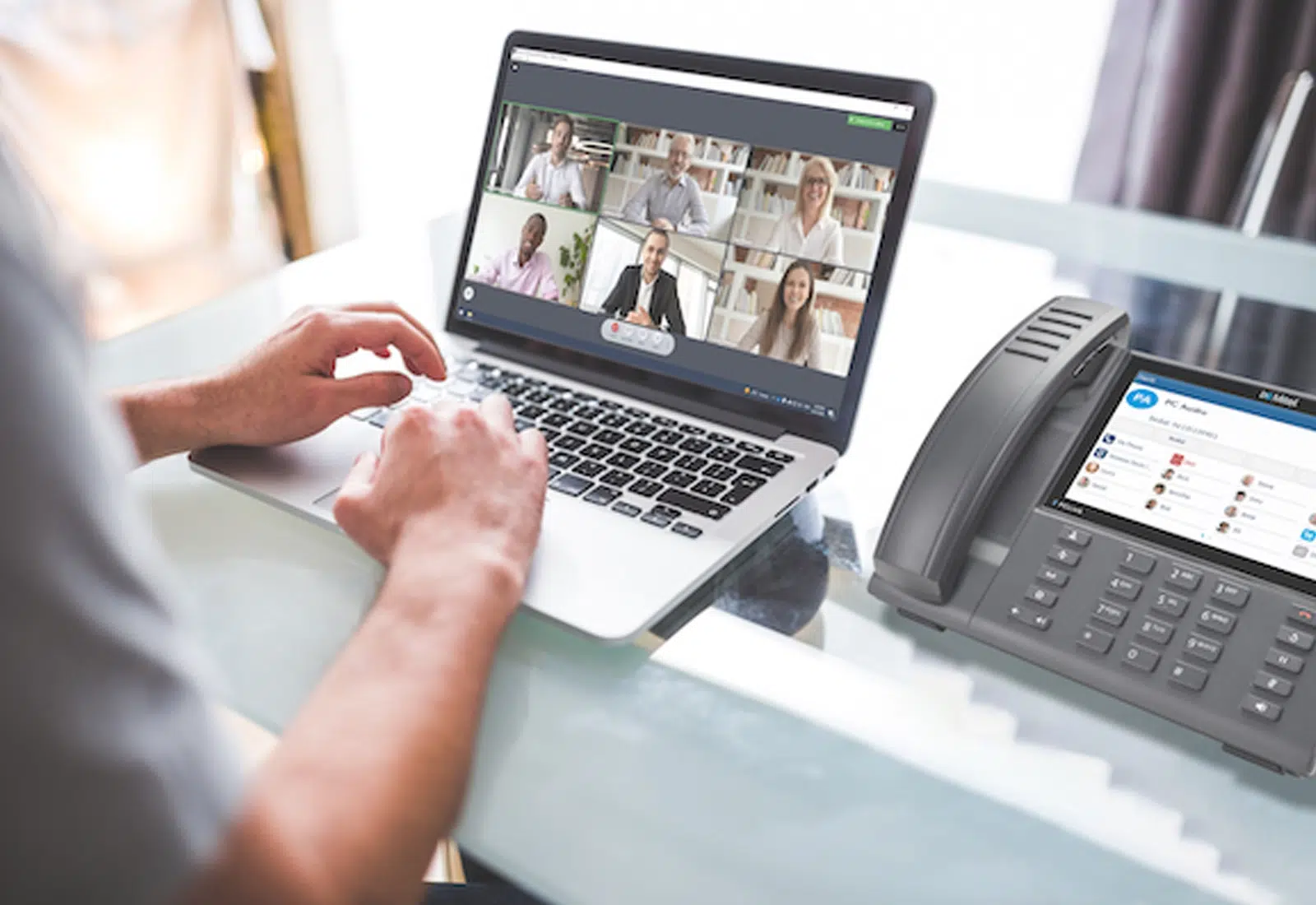 A close up of an employee using a laptop for a conference call over VoIP.  On the laptop screen are six other people on the video call. Next to the laptop is a Mitel IP phone.