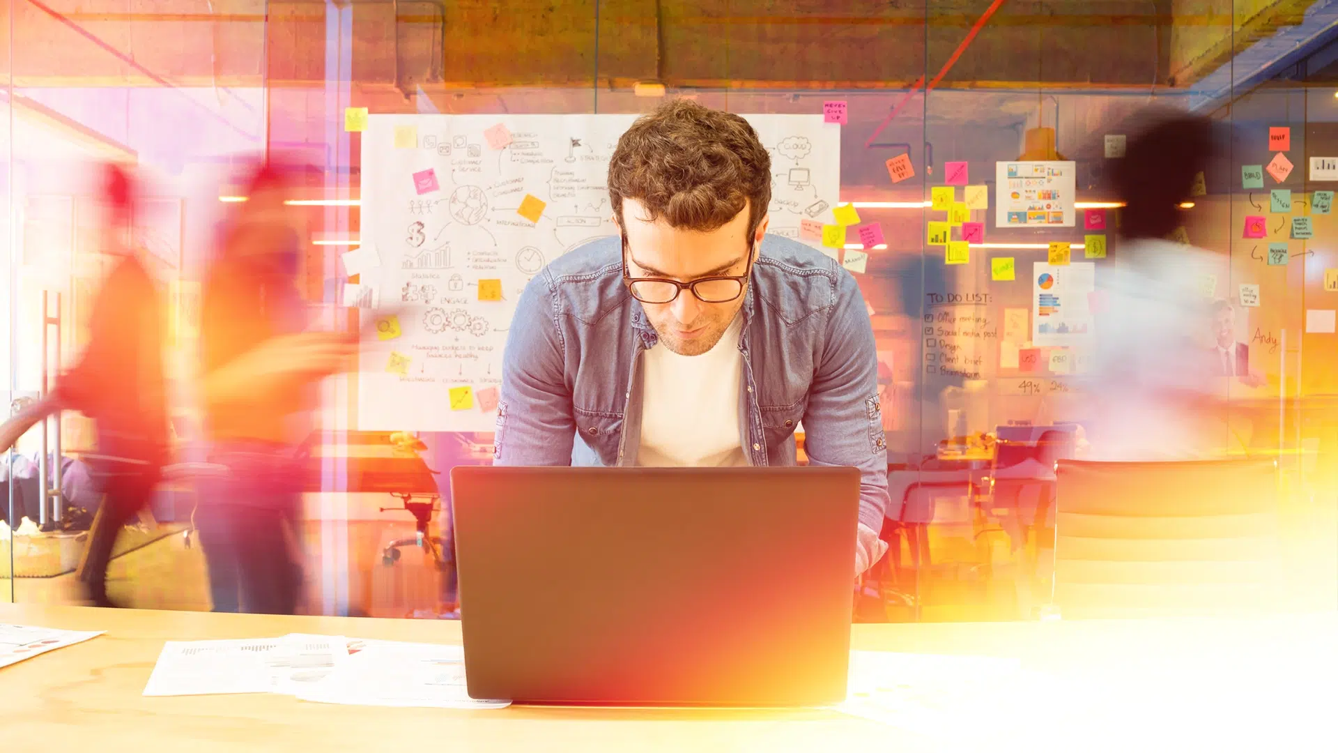 A Telexperts employee works on VoIP systems at a laptop. Behind him is an office setting with a post board and two other workers, all blured out of focus.